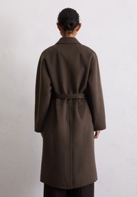 Person mit einem Haarknoten, die einen langen, dunkelbraunen mantle mit Gürtel trägt und mit dem Rücken zur Kamera vor einer schlichten, hellgrauen Wand steht.