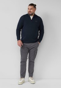 Navy gebreid vest met rits en lange mouwen, gecombineerd met grijze cargobroek met zakken en witte sneakers met crème accenten.