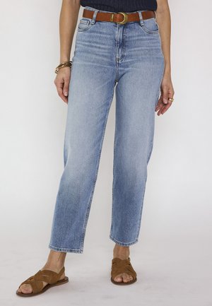 Personne portant un jean taille haute bleu clair, ceinture marron avec boucle dorée, haut noir, bracelets dorés, bague et sandales plates marron.