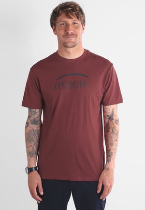 T-shirt en coton bordeaux avec un logo "OXBOW" noir, col rond, manches courtes et coupe droite. Comprend des bras tatoués visibles sur l'image.