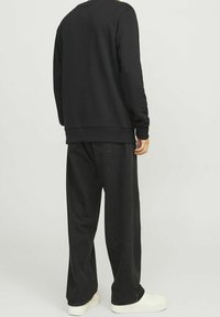 Una persona sta in piedi con le spalle alla telecamera, indossando una felpa nera e pantaloni neri larghi, con delle sneakers bianche su uno sfondo chiaro.