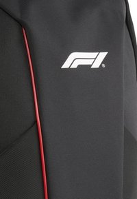 Sorte struktureret rygsæk med en rød kantlinje, der har et hvidt Formel 1-logo på siden. Lidt geometrisk design.