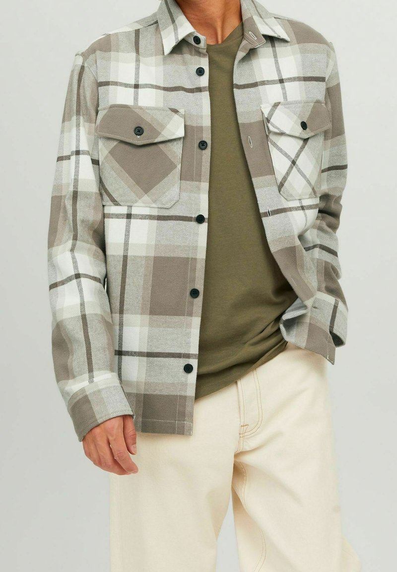 Mies, joka on pukeutunut vaaleanbeigeen ja valkoiseen ruudulliseen napilliseen paitaan oliivivihreän t-paidan päällä, beigeihin housuihin, seisoo tasaisen taustan edessä.