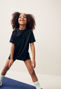 Czarny sportowy t-shirt i szorty, w luźnym kroju. Białe skarpetki w zielone paski. Dziecko z kręconymi włosami radośnie pozujące.