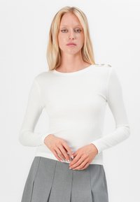 Long-sleeve white ribbed top with a round neckline and decorative buttons on the left shoulder, paired with a gray pleated skirt.