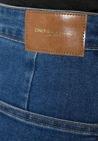Jeans de ganga azul apresentam um remendo de couro castanho na cintura com texto em relevo, detalhes dourados e costuras contrastantes ao longo das costuras.