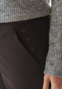 Zwarte broek van gladde stof, voorzien van een diagonale zak met drie zwarte knopen, gecombineerd met een lichtgrijze gestructureerde trui.