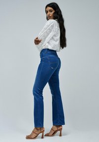 A woman with long black hair poses sideways, wearing a white blouse and blue jeans with heeled sandals, against a neutral background.