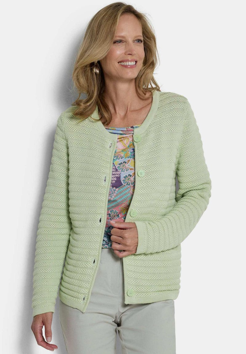 Femme souriante portant un cardigan boutonné texturé vert clair sur un haut à motif floral et un pantalon pâle, devant un fond neutre.