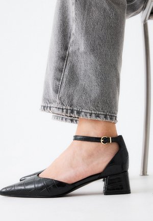 Zapatos de charol negro de tacón bajo con punta afilada y tira en el tobillo, con textura de cocodrilo y un detalle en hebilla dorada.