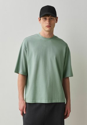 Ung mand iført en løs lysegrøn T-shirt, sort kasket og mørke bukser, stående mod en ensartet lysegrå baggrund.