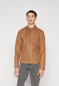 Veste en suède marron avec un col montant, fermeture éclair à l'avant et deux poches latérales. Le tissu a une finition douce et texturée. Portée avec un jean gris.