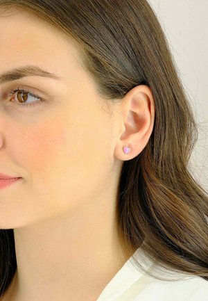 Orecchini a forma di cuore in tono oro, con una finitura in smalto rosa pastello, impreziositi da un piccolo accento di gemma, indossati all'orecchio di un modello.