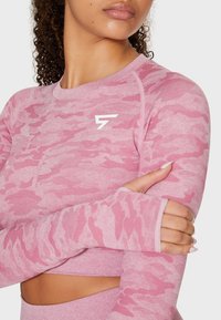 Femme portant un crop top à manches longues camouflage rose avec des trous pour les pouces et un petit logo blanc sur la poitrine.