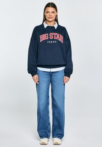 Námořnická fleecová mikina s grafikou "BIG STAR JEANS", v kombinaci s světle modrou košilí s límečkem a modrými džínami s širokými nohavicemi, krémové tenisky.