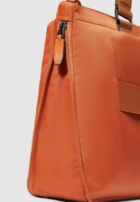 Oranje stoffen handtas met een getextureerd oppervlak, ritssluiting aan de bovenkant en een brede, gestructureerde vorm. Voorzien van een klein leren accent bij de rits.