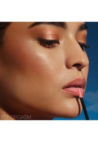 Lipgloss in een zachte roze tint, aangebracht met een applicator. De huid van het model heeft een onberispelijke, stralende finish, met koperkleurige oogmake-up.