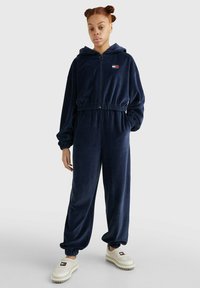 Navy velvet jumpsuit met een ritssluiting aan de voorkant, capuchon en elastische tailleband. Kenmerkt ribgebreide boorden en een klein merklabel op de borst.