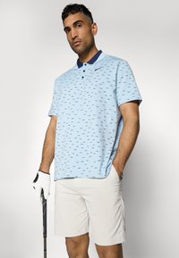 Light blue polo shirt with a patterned design, featuring a dark collar, paired with beige shorts. The model holds a golf club and wears a white glove.