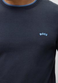 Jersey azul marino con cuello redondo acanalado, que presenta un logo "BOSS" en azul claro en el lado izquierdo del pecho. Textura suave y diseño ajustado.