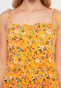 Vestido amarelo florido com alças finas, decote franzido, apresentando flores e folhas multicoloridas em tecido suave.