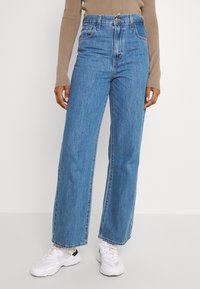 Person klädd i högmidjade, raka blåjeans, beige långärmad topp och vita sneakers mot en enkel vit bakgrund.