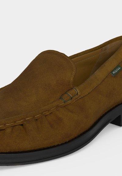 Καφέ σουέτ μοκασίνι slip-on με μαύρη σόλα, λεπτομερή ραφές και μικρή υφασμάτινη ετικέτα στο πλάι.