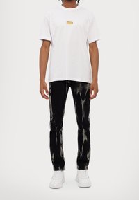 Vit bomullströja med en liten guldig logotyp på bröstet, matchad med svarta jeans med ett blekat mönster och vita sneakers.