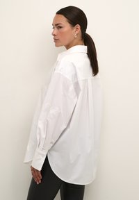 Camicia bianca con maniche lunghe, motivo a farfalla ricamato in modo sottile sulle maniche, orlo arrotondato e polsini con bottoni. Modello di spalle rispetto alla fotocamera.