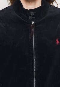 Black corduroy jacket with a high collar, two buttons, and a front zipper. Features a small red logo on the chest area.