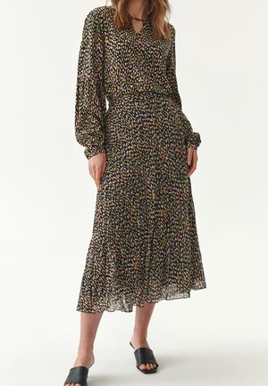 Vrouw die een midi-jurk met lange mouwen en een meerkleurig patroon draagt, met zwarte open-toe instapschoenen, staand tegen een effen witte achtergrond.