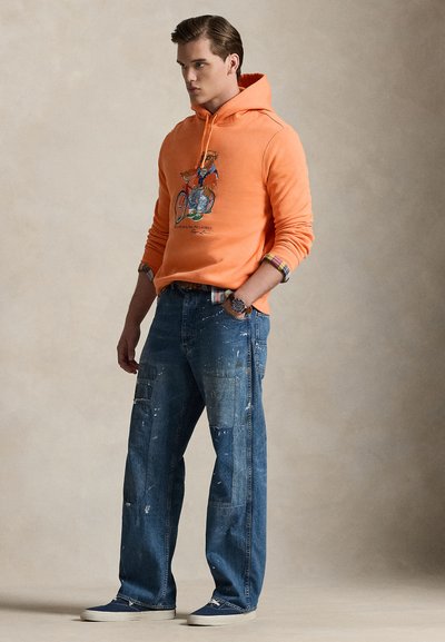Πορτοκαλί φούτερ με κουκούλα με γραφικό ποδηλάτου, συνδυασμένο με μπλε τζιν παντελόνι με φθορές, πολλαπλές τσέπες και πιτσιλιές μπογιάς. Λευκά αθλητικά παπούτσια.
