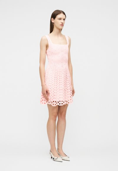 Γυναίκα που στέκεται φορώντας ένα αμάνικο, απαλό ροζ φόρεμα με λέιζερ και τετράγωνο ντεκολτέ, μαζί με λευκά παπούτσια με χαμηλό τακούνι και μυτερή μύτη.