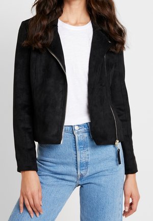 Kvinde iført en sort ruskindjakke med diagonalt lynlås over en hvid t-shirt og lyseblå højhælede jeans.