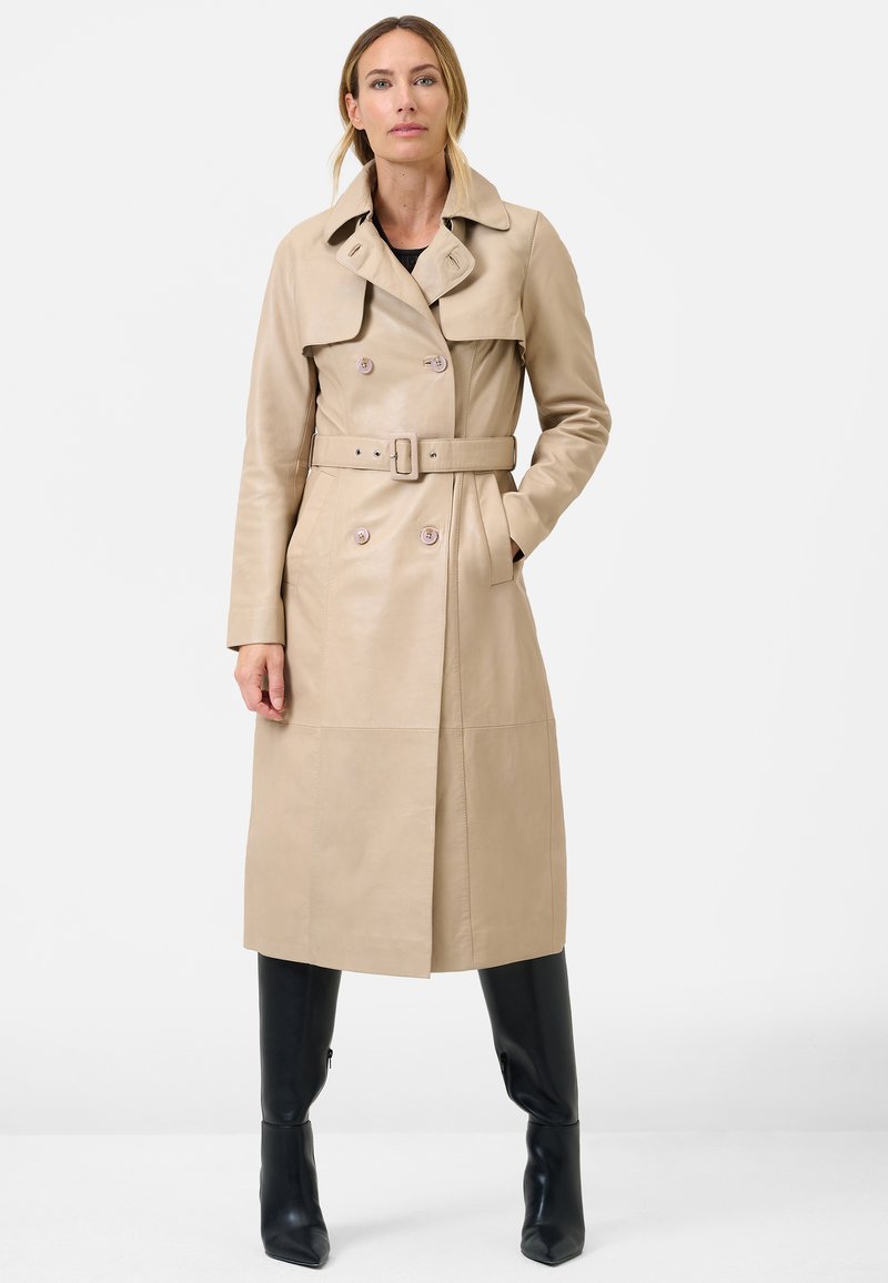 Beige leather trench coat with double-breasted buttons, wide collar, and waist belt. Smooth texture, knee-length design. Black fitted pants.