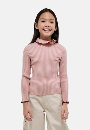 Fille souriante portant un pull rose côtelé avec un col et des poignets à volants, ainsi qu'un pantalon beige clair avec des détails brodés sur les côtés.