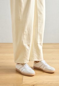 Sneaker bianco con pannelli in suede e rete, dotato di suola in gomma e design con lacci. Abbinato a pantaloni a gamba larga beige chiaro.