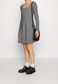 Schwarz-weißer karierter Kleid mit langen Ärmeln und quadratischem Ausschnitt. Kombiniert mit schwarzen Plateauschuhen und weißen Rüschenstrümpfen.