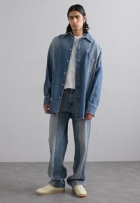 Denim srajca in hlače v različnih odtenkih modre, z ohlapnim krojem. Bela mrežasta majica pod njimi, v kombinaciji s svetlimi supergami.