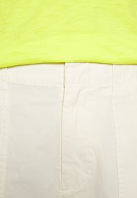Chemise à manches courtes jaune fluo superposée sur un pantalon couleur crème. Le pantalon présente une taille plate avec des détails de couture subtils.