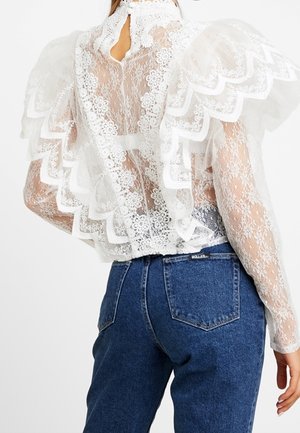 Blouse en dentelle blanche transparente avec motifs floraux, grands volants festonnés, col montant, associée à un jean en denim bleu.