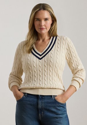 Femme aux longs cheveux blonds portant un pull crème à col en V en maille torsadée avec des rayures bleu marine et un jean bleu, mains dans les poches, fond neutre.