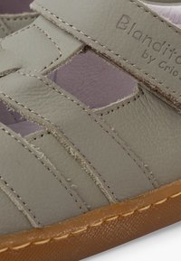 Gros plan sur des lanières de sandale en cuir gris avec des coutures visibles et une semelle en caoutchouc marron, montrant le nom de la marque « Blandito » embossé.
