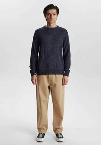 Ciemnoniebieski sweter o teksturowanej powierzchni z okrągłym dekoltem, zestawiony z beżowymi spodniami typu chinos o szerokich nogawkach i czarnymi sneakersami. Prosty design i luźny krój.