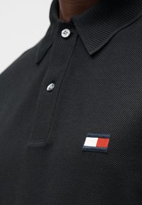 Gros plan d'un polo noir texturé avec deux boutons et un petit logo rectangulaire rouge, blanc et marine sur la poitrine.