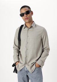 Camisa de botones de manga larga gris claro con cuello, combinada con jeans azules. Se incluyen gafas de sol oscuras y una bolsa de asa negra.