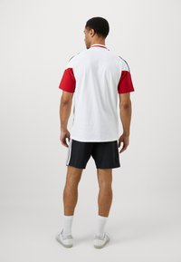 T-shirt branca com mangas vermelhas, detalhes pretos e colarinho, combinada com calções pretos com riscas brancas nas laterais.