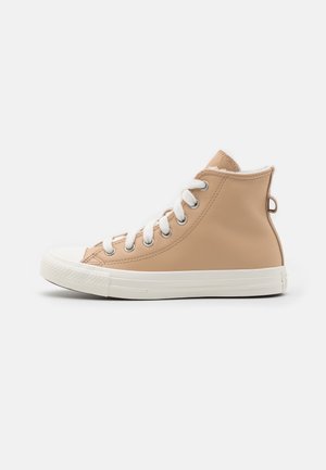 Beige hoge sneaker met witte veters, metalen oogjes, rubberen neuskap en witte rubberen zool op een eenvoudige lichte achtergrond.