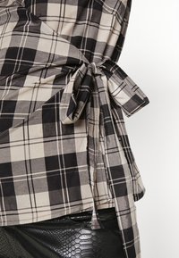 Camisa xadrez preta e bege com detalhe de laço na cintura. O tecido parece suave com uma textura lisa e mangas curtas.