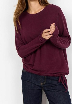 Femme portant un pull à manches longues violet foncé avec un détail de nœud sur le côté, associé à un jean bleu foncé, debout devant un fond uni.